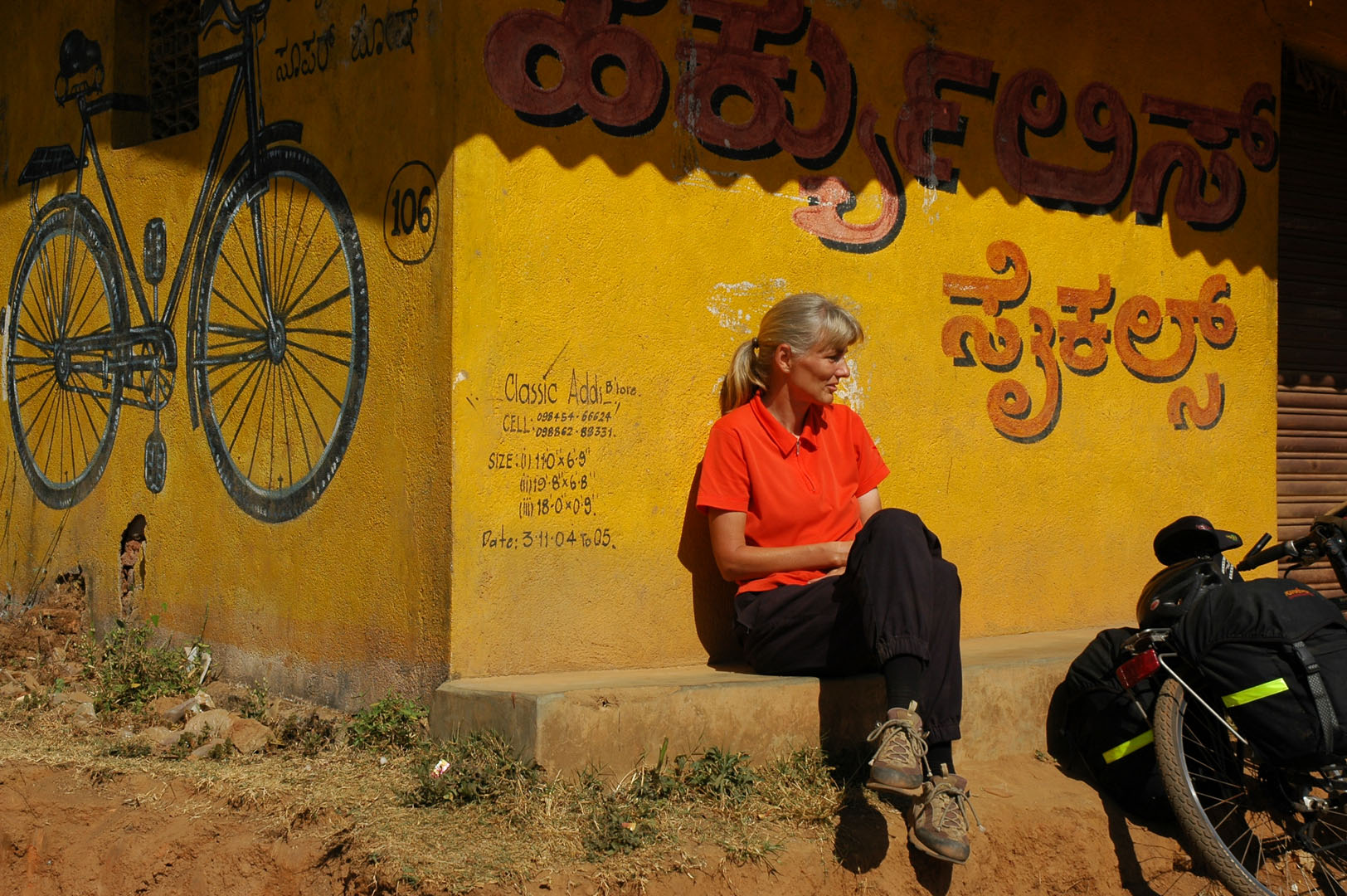 A western cyclist leans against a building with an Indian bicycle painted on it during a bicycle tour of South India.