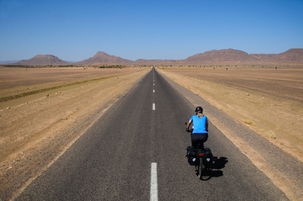 A blue touring cyclist pedals towards brown hills in the Morroccan desert.